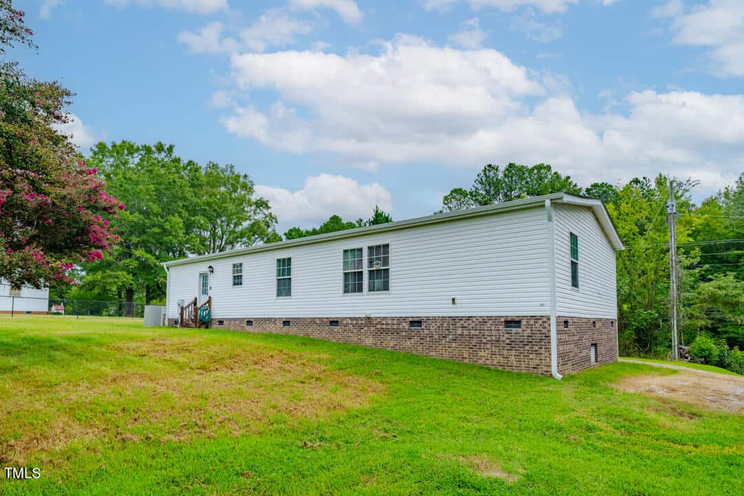 A single-story white manufactured home with a brick foundation sits on a grassy lawn. A small set of steps leads to the entrance, and the background features trees and a cloudy sky.