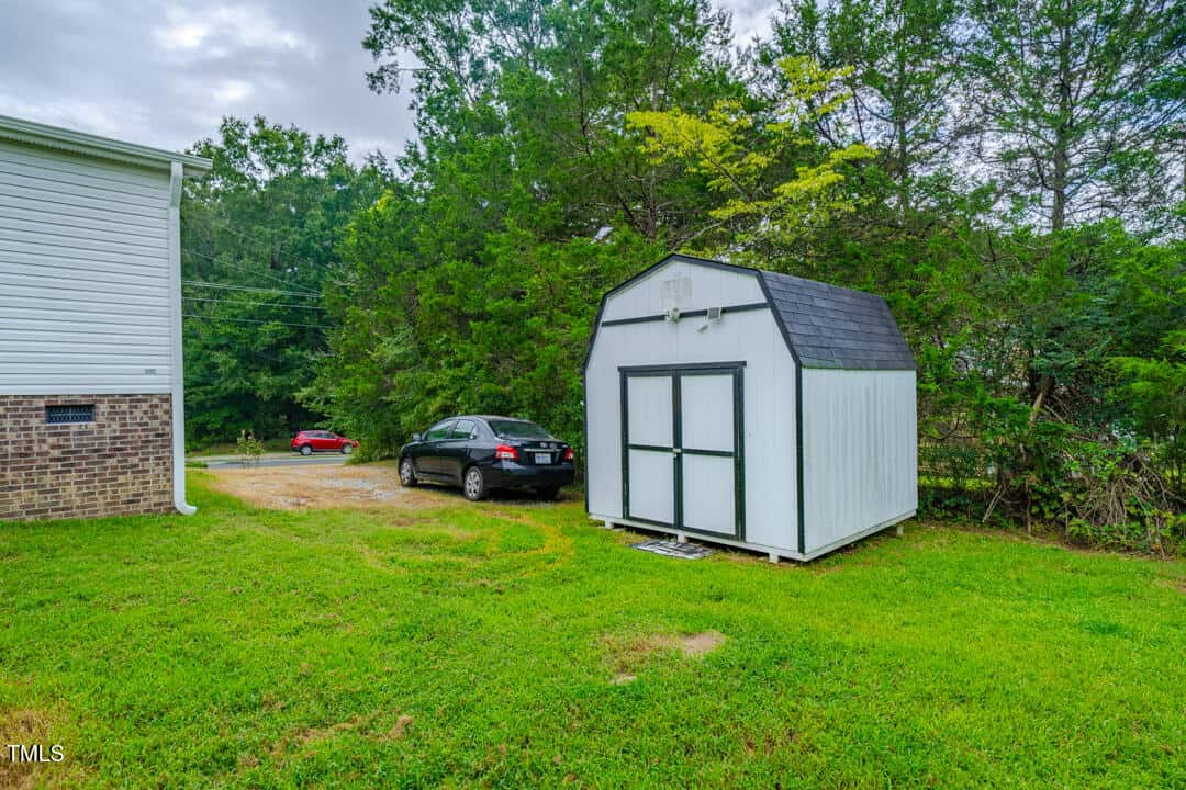 A white shed with a black roof stands in a grassy backyard next to a brick building. Two cars are parked beside the shed, and tall green trees line the background.
