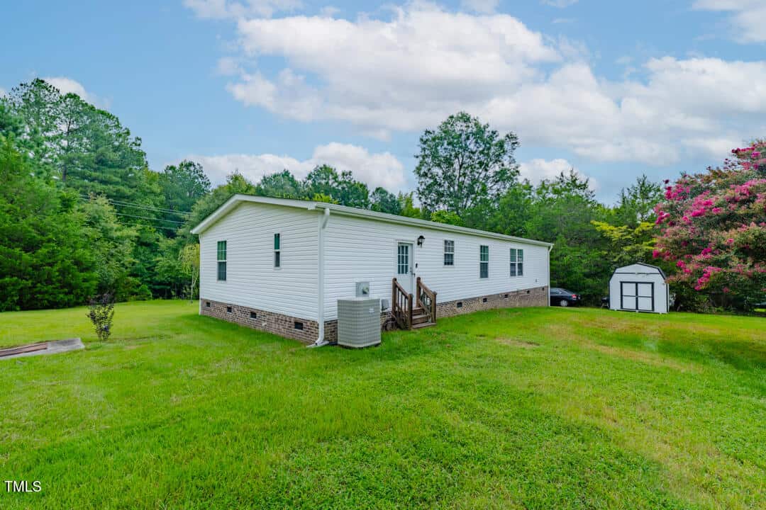 Single-story white home with a brick foundation surrounded by lush green lawn and tall trees. A small shed sits nearby. The sky is partly cloudy.