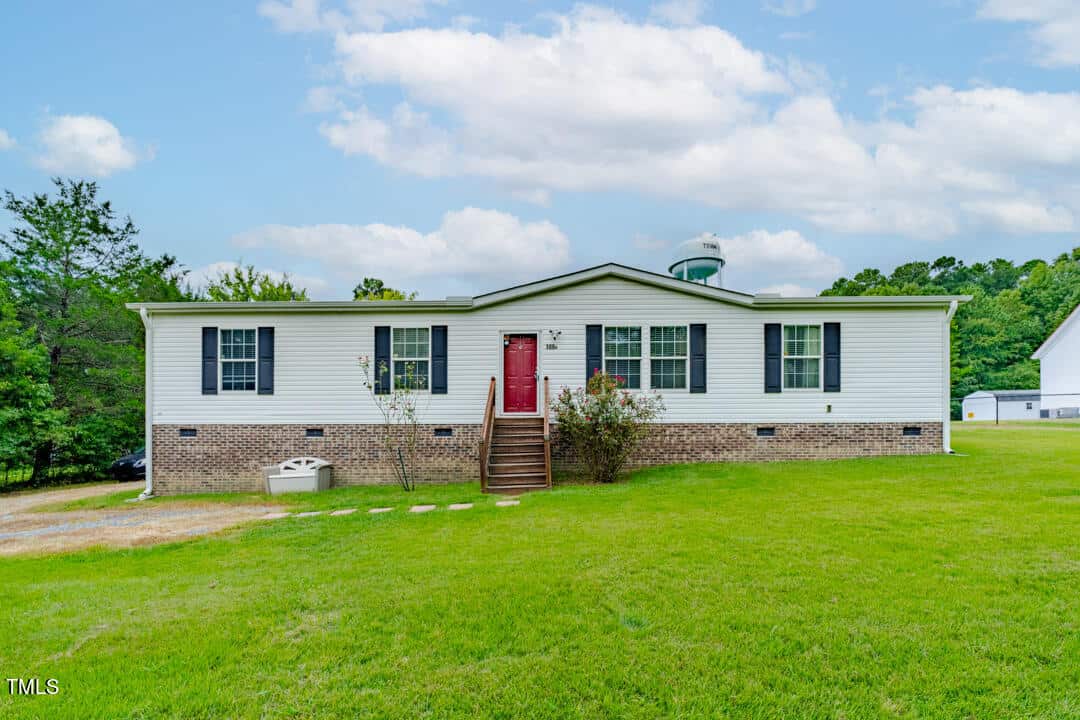 A charming single-story house with a red door and white siding sits on a grassy lawn at 109a Old Nc 75. Steps lead to its entrance, under a partly cloudy sky. Bushes and trees stem around the property, while a neighboring house peeks from the background.