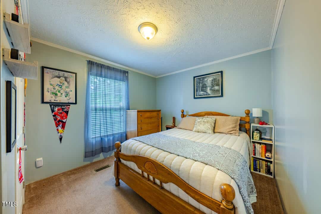 A cozy bedroom with a wooden bed, light blue walls, and a carpeted floor. There's a dresser and bookshelf beside the bed, two framed pictures on the walls, and a window with sheer curtains allowing natural light to enter.