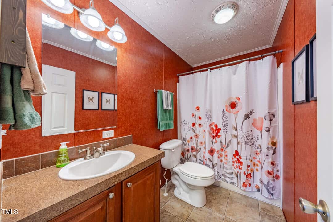 A bathroom with red-orange walls, a sink with a brown countertop, and a mirror above it. Towels hang beside the mirror. The shower curtain features a floral pattern in red and gray. The floor is tiled, and there are lights above the mirror.