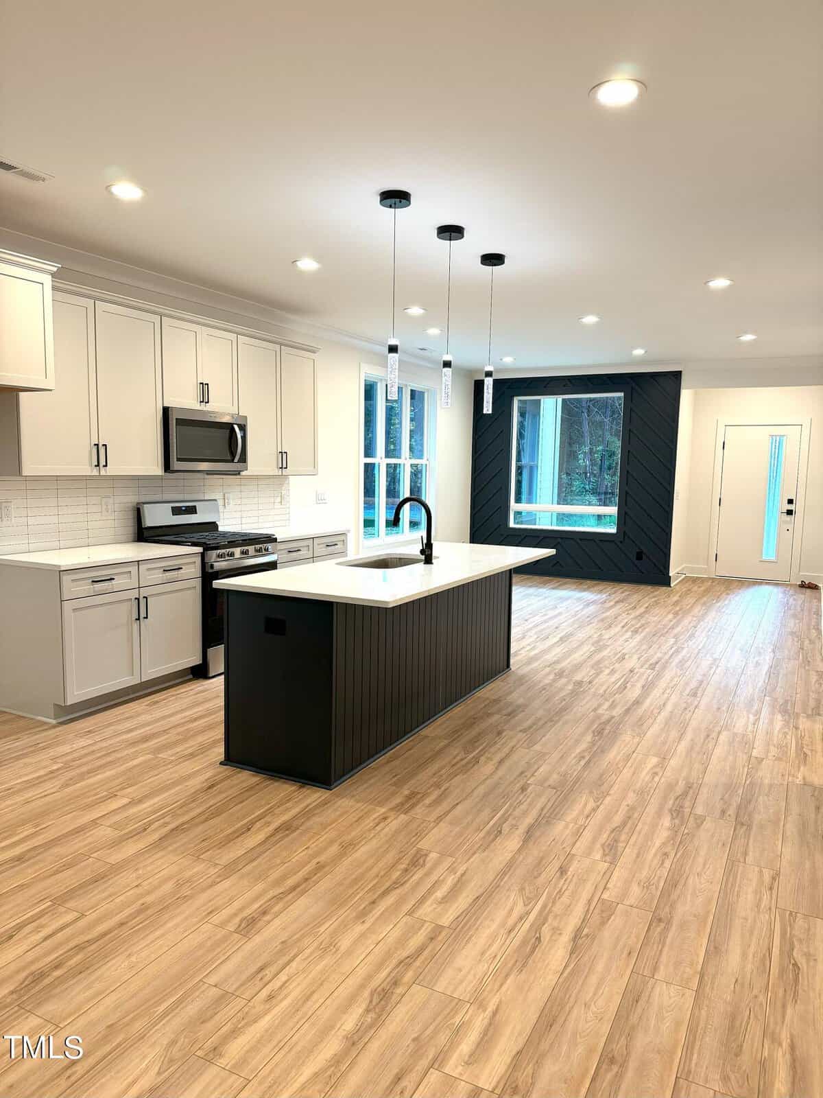 Modern kitchen with white cabinets, stainless steel appliances, a black island with a sink, pendant lights above, light wood floors, large windows, and a glass door leading outside.