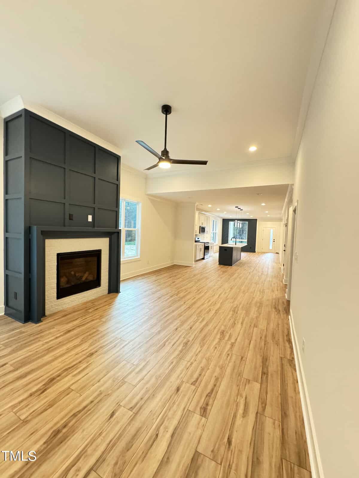 Spacious modern living area with light wood flooring, a dark accent wall featuring a fireplace, ceiling fan, large windows, and an open layout leading to a kitchen with an island and recessed lighting.