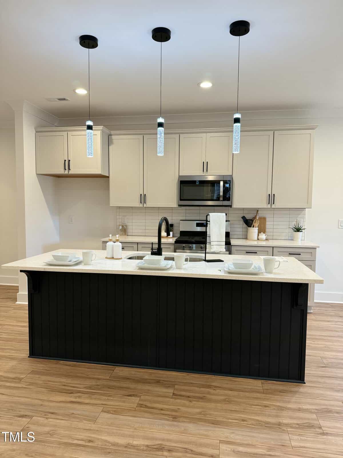 Modern kitchen with light-colored cabinets, stainless steel appliances, a black island with a white countertop, three pendant lights, and wood flooring. White dishes and cups are arranged on the island.