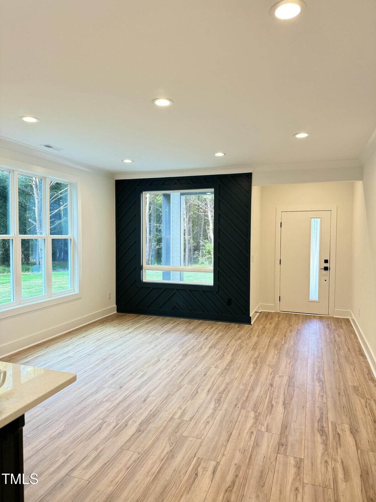 Bright modern living room with large windows, light wood flooring, recessed ceiling lights, a black accent wall with geometric pattern, and a white front door with a glass panel. Forested area visible outside.