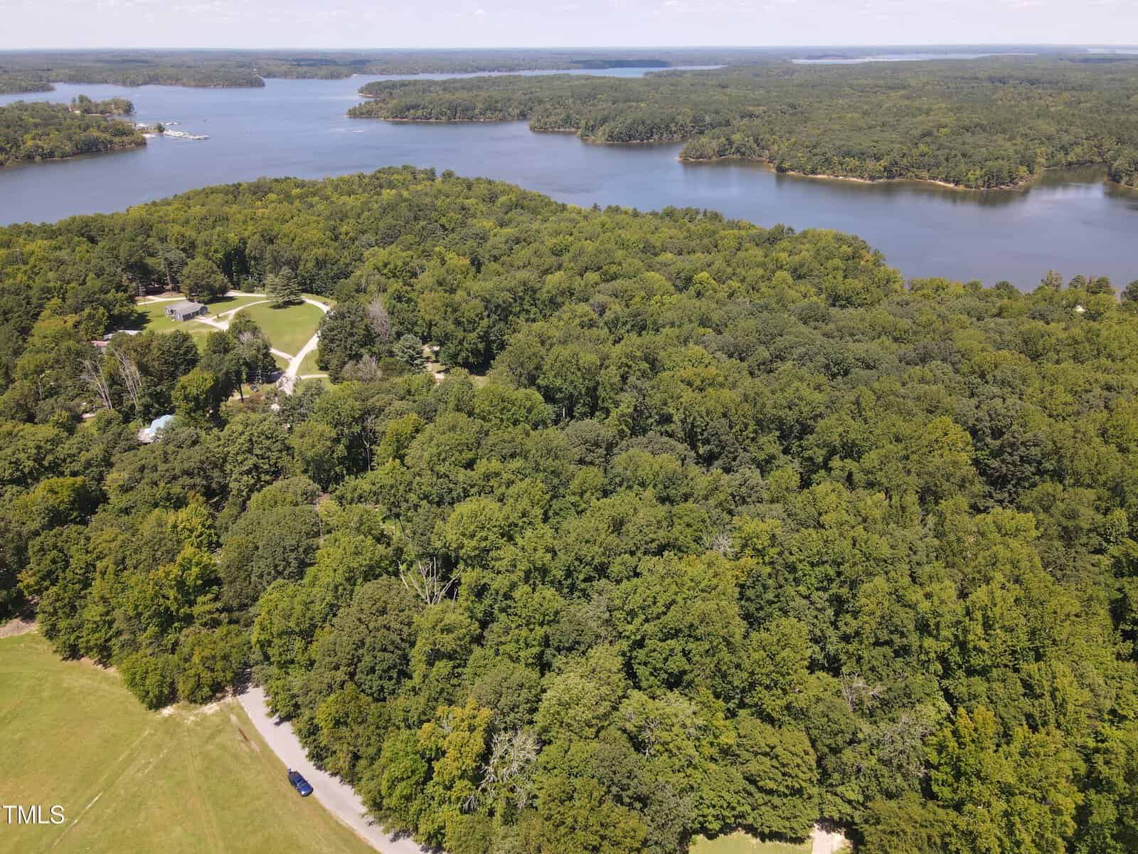 Aerial view of a large, lush green forest surrounding a winding lake. The scene includes a few scattered buildings, open grassy areas, and a narrow road cutting through the landscape. Several peninsulas extend into the calm waters of the lake.