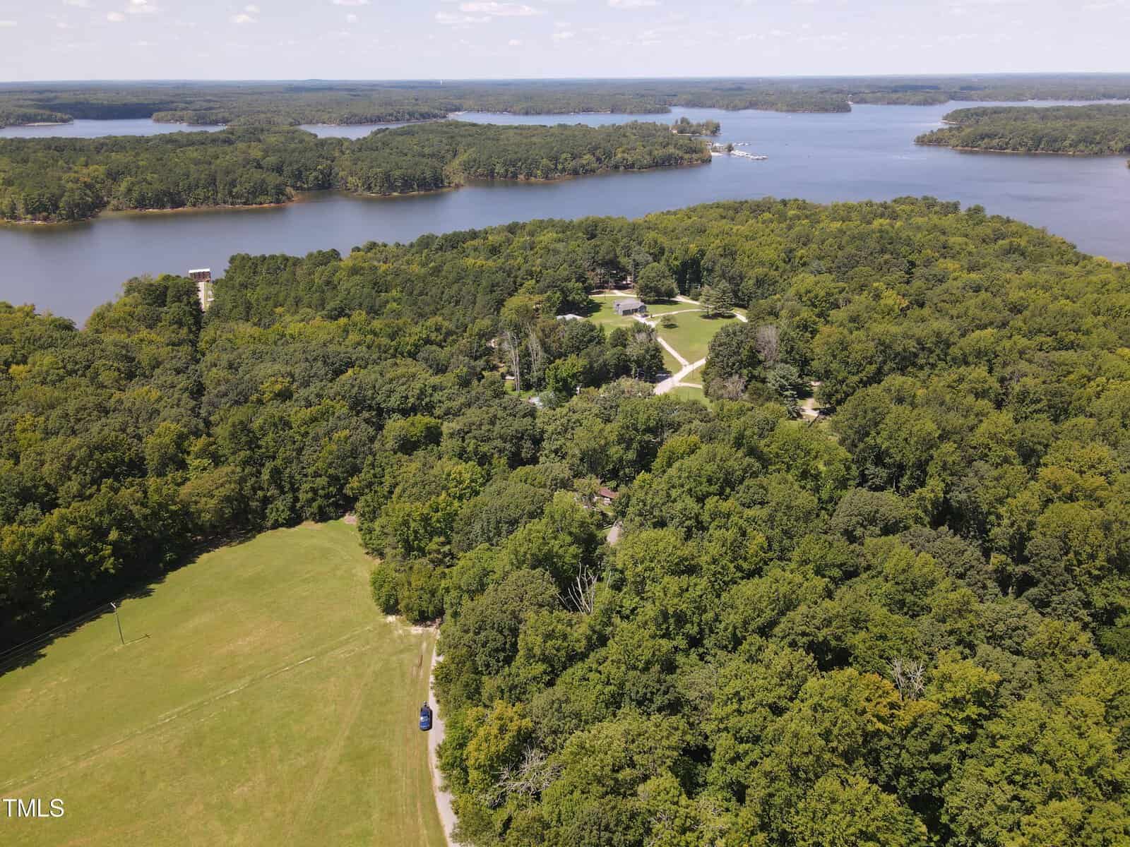 Aerial view of a large lake surrounded by dense green forests. A winding road cuts through the trees leading to a clearing with several buildings. The lake curves around the wooded area, with scattered islands visible. Jeep parked in the open field in the foreground.