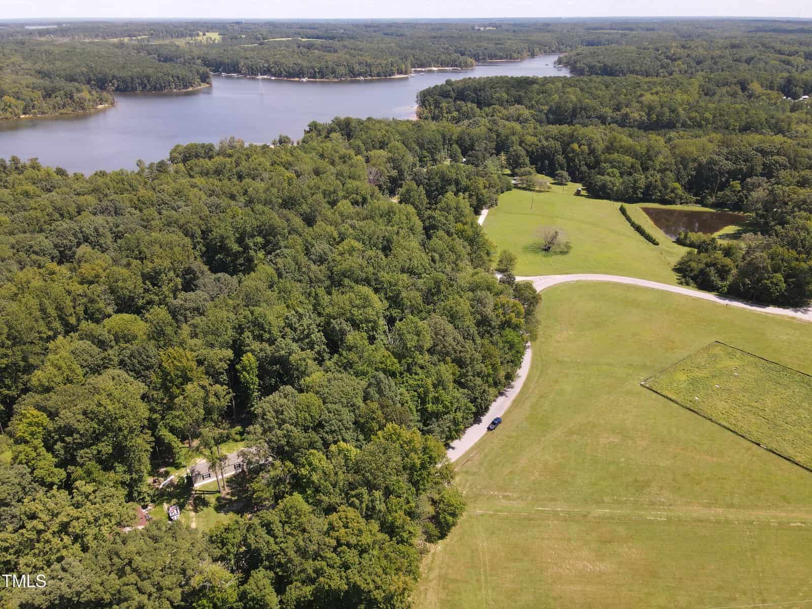 Aerial view of a scenic landscape featuring a large, winding river surrounded by dense forests and open fields. A curved road cuts through the greenery, with a few scattered buildings visible among the trees. The image portrays a serene and natural environment.