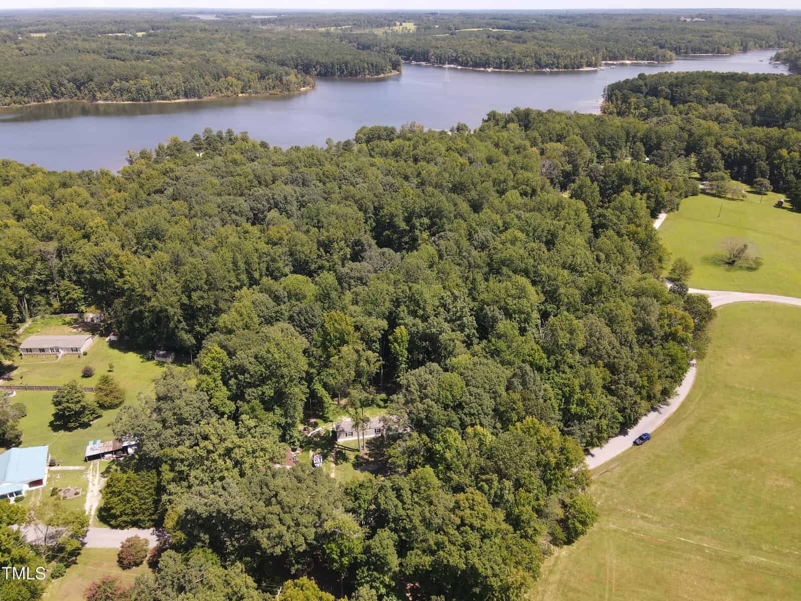 Aerial view of a forested area with dense greenery surrounding a few scattered houses. A winding road cuts through the landscape, leading towards a large lake in the background. The scene is peaceful and lush, showcasing the natural beauty of the region.