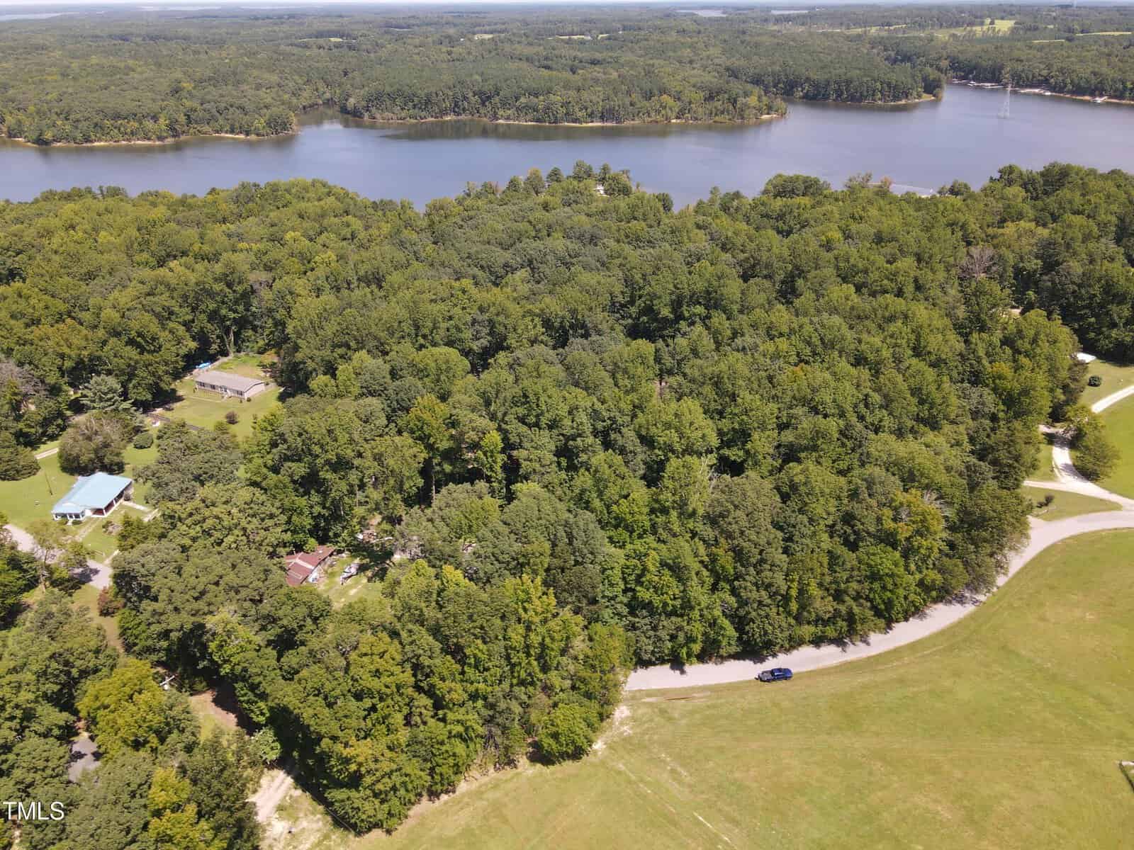 An aerial view of a densely wooded area beside a large lake. A few houses are visible among the trees, with a winding road cutting through the forest. The lake's shoreline is lined with trees, and a large grassy area is in the foreground.