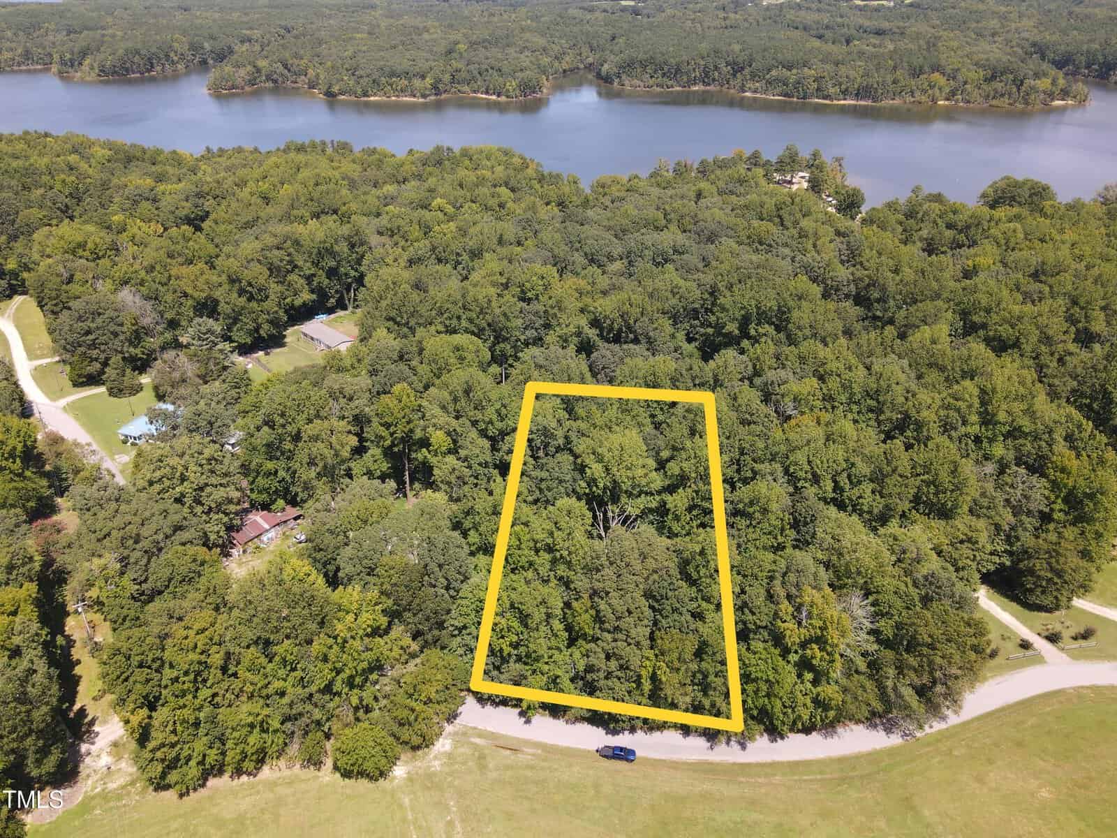 Aerial view of a dense forested area with a rectangular plot outlined in yellow near 000 Intake Lane, Henderson. Nearby, a road winds past a blue vehicle, and in the background, there is a large body of water surrounded by more trees. A few structures are visible among the trees.