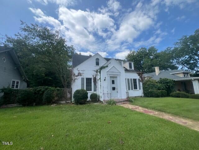 A white, two-story house with an A-frame roof and a small front porch. The house is surrounded by a grassy lawn, bushes, and trees on both sides. The sky above is partly cloudy.