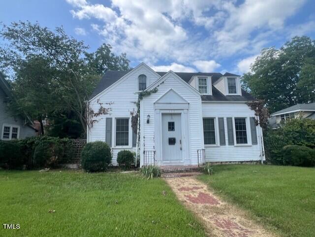 A white, two-story house at 109 W Jennette Avenue in Henderson boasts a gabled roof and two dormer windows. The front entrance features a small overhang, flanked by tall bushes. The front yard consists of green grass and a narrow brick pathway leading to the door. The sky above is partly cloudy.