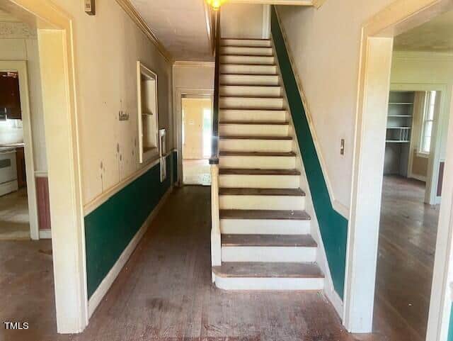 A dimly lit hallway with wooden flooring leads to a staircase with green and white walls. The hallway extends to adjoining rooms on both sides. The area appears somewhat aged, with visible dirt on the floor and walls. The staircase has narrow steps and a wooden banister.