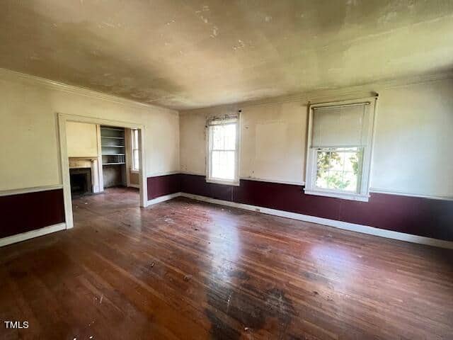 A spacious, vacant room with worn hardwood floors and partially cracked, off-white walls. The room features two windows with white trim, and an open doorway leading to another room with built-in shelves and a fireplace. Some debris is scattered on the floor.