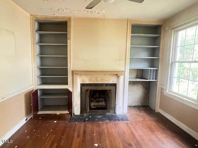 A room with beige walls and hardwood flooring featuring an empty fireplace with an ornate mantel. There are built-in shelves on both sides of the fireplace, some with missing or unpainted sections. Paint is peeling from the ceiling, and a large window offers natural light.