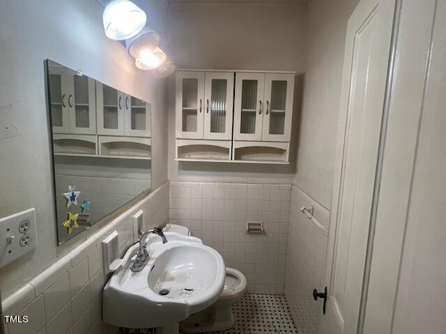 A small bathroom featuring a wall-mounted sink, a large mirror above it, and a toilet. Overhead, there's a light fixture with multiple bulbs and a cabinet with glass-paneled doors for storage. The floor is tiled in a mosaic pattern.