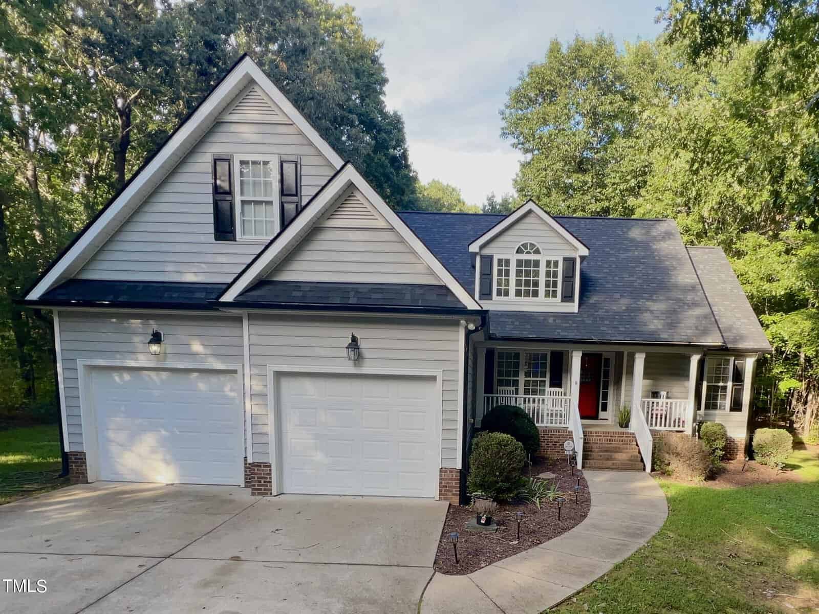 Nestled on 4143 Geneva Drive in Franklinton, this two-story house boasts gray siding, a dark roof, and two white garage doors. A vibrant red door with a cozy porch and white railings is framed by shrubs. Beyond, trees and a lush yard complete the charming backdrop.