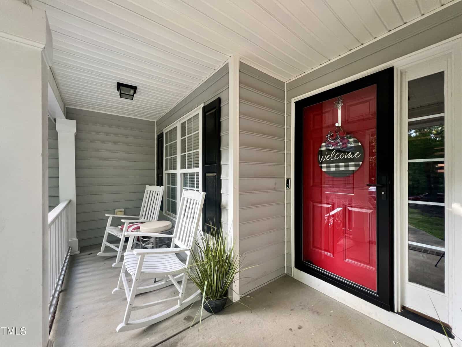 A cozy porch with gray siding, featuring two white rocking chairs and a small table between them. A welcoming red door with a decorative sign hangs nearby. A potted plant adds greenery to the space.
