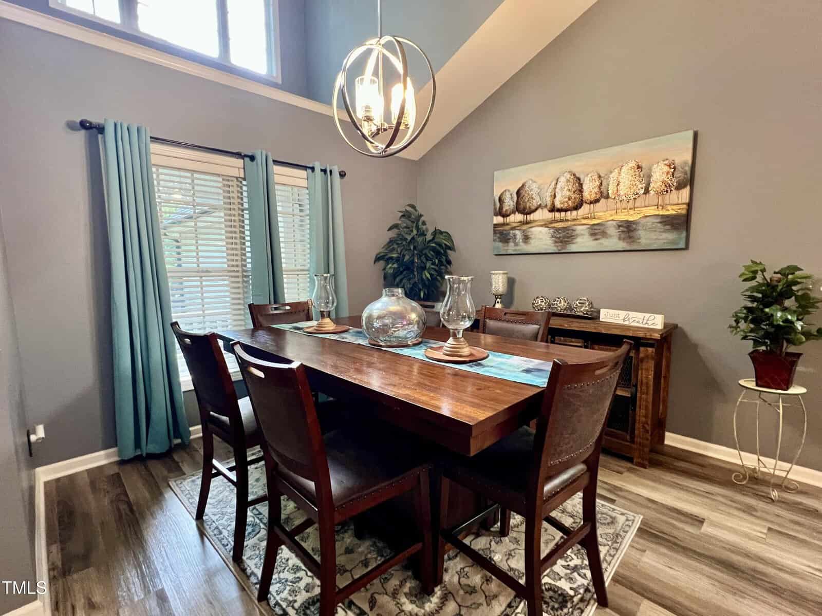 A dining room with a wooden table set for six, featuring decorative items. Blue curtains adorn the window, and a large nature-themed painting hangs on the gray wall. A modern chandelier lights the room, which has wood flooring and plants.