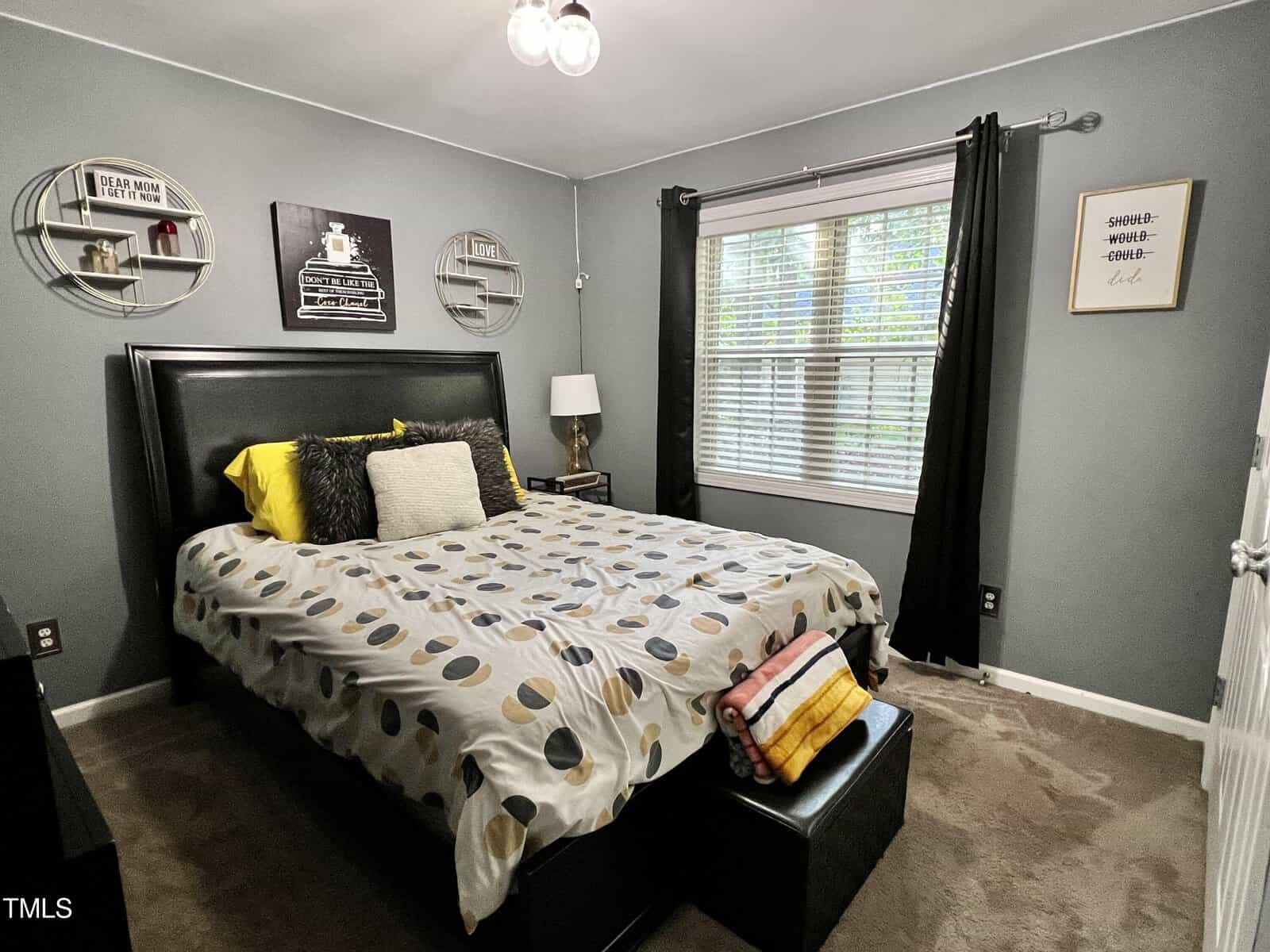 A cozy bedroom with a large bed featuring polka dot bedding. The room has gray walls, a window with black curtains, a lamp on a bedside table, and decorative wall art. A bench is positioned at the foot of the bed.