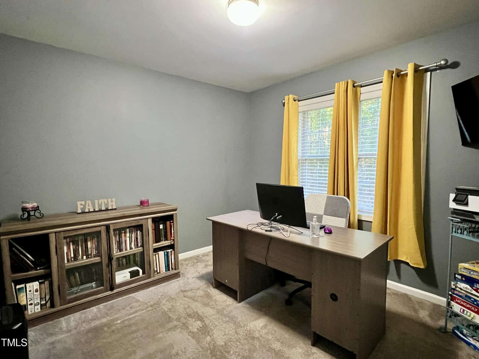 A home office features a wooden desk with a computer monitor, an office chair, and a bookshelf filled with books and decor items. Bright yellow curtains hang by the window, illuminating the gray walls and carpeted floor.