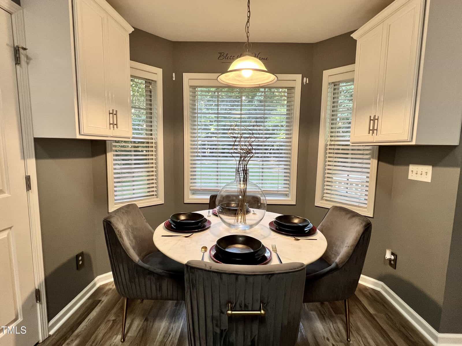 A cozy dining area with a round marble table surrounded by four gray upholstered chairs. The table is set with black plates and bowls. A large window with blinds overlooks the outside. Cabinets are on either side, and a pendant light hangs overhead.