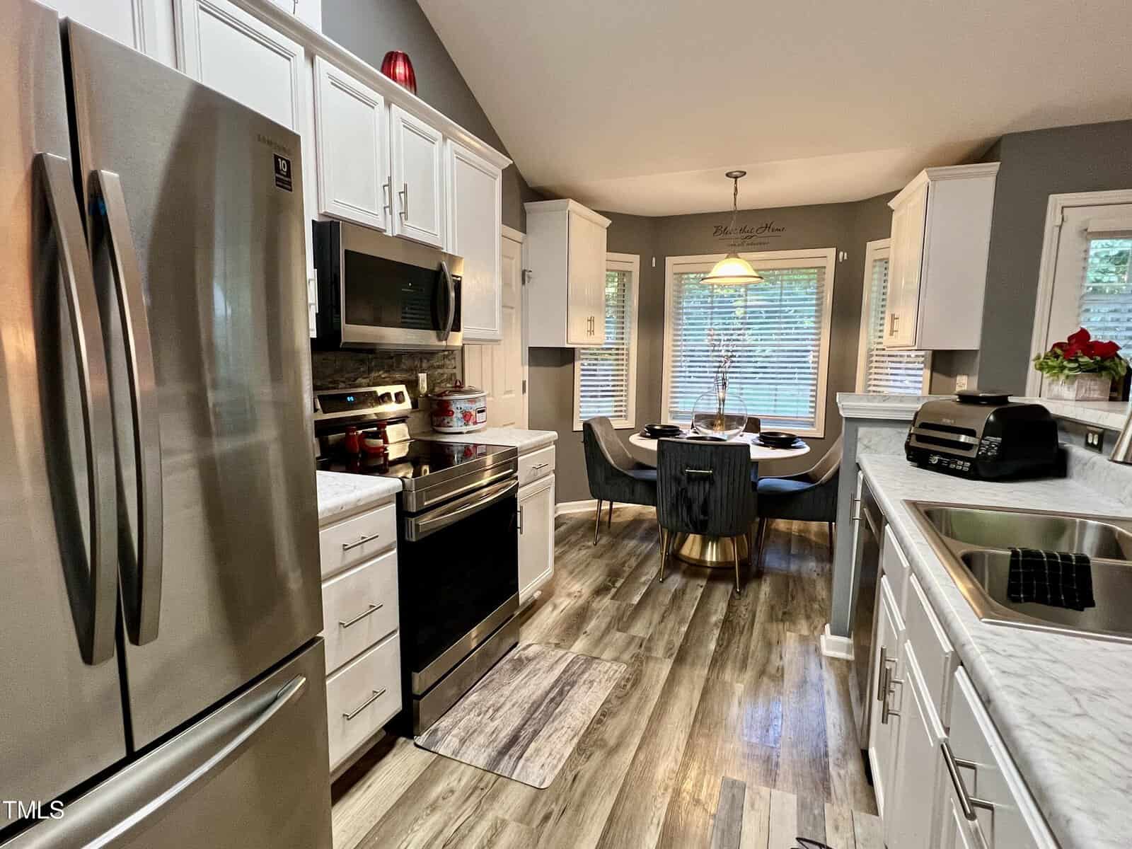 Modern kitchen with stainless steel appliances, white cabinets, and marble countertops. A small dining area is visible with a round table and chairs by the window. Natural light illuminates the space, highlighting the laminate wood flooring.