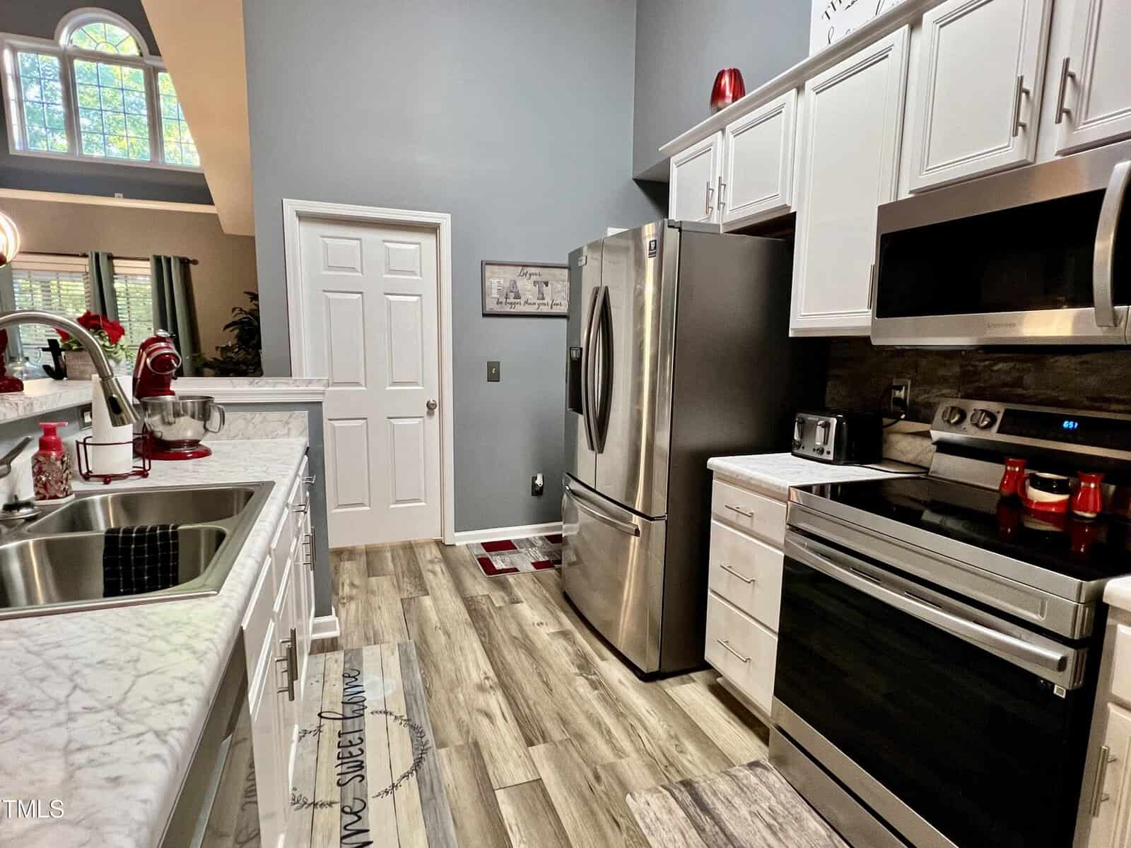 A modern kitchen with marble countertops, a stainless steel refrigerator, and an electric stove. Red accents include a mixer and kettle. Light-colored wooden floors and white cabinets add a fresh, clean look. A door and arched window are visible.