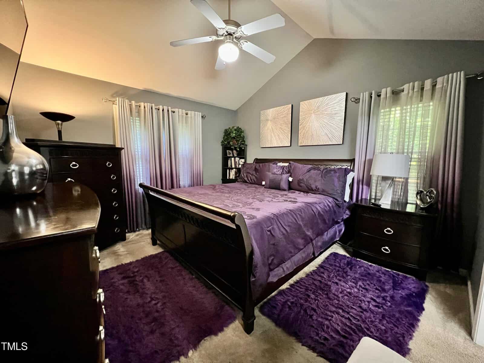 A bedroom with a dark wooden bed and purple bedding. Two abstract paintings hang above the bed. There are matching end tables with lamps, a tall dresser, a ceiling fan, and curtains on the windows. Purple rugs cover parts of the carpeted floor.