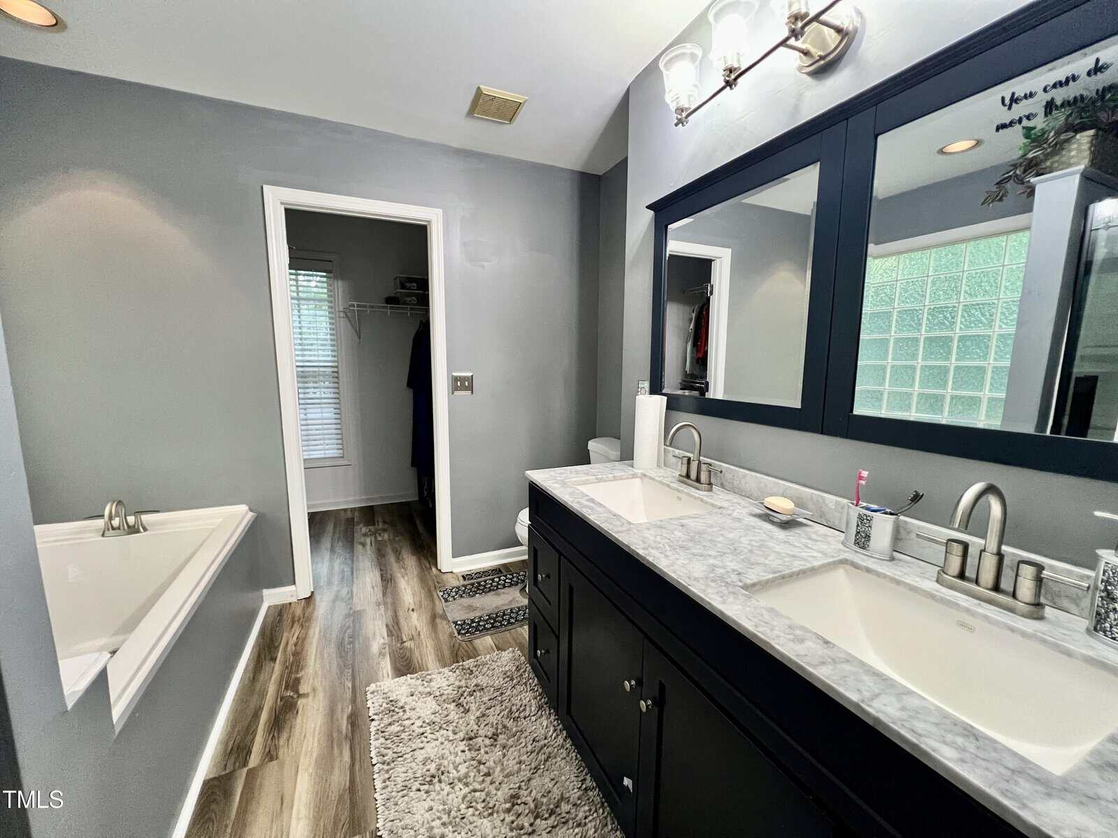 Modern bathroom with a dark wood vanity, dual sinks, and a large mirror. A bathtub is on the left, and the room features gray walls and wood flooring. A doorway leads to a closet with shelves. A textured rug is in front of the vanity.