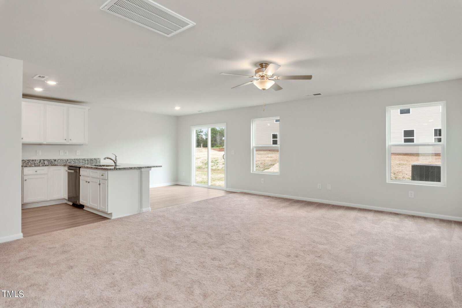 A spacious, bright, unfurnished living room with light beige carpet, an open layout, and a ceiling fan. In the background, a modern kitchen with white cabinets and a granite countertop. Large windows offer natural light and a view of the yard.