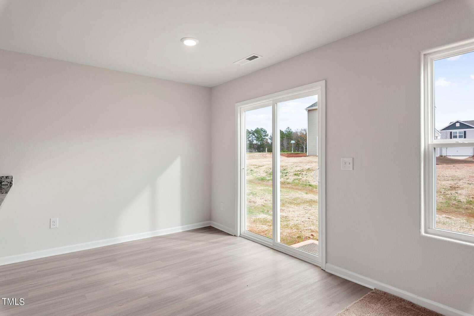 Bright room with light wood flooring, a sliding glass door, and a large window offering a view of an open field and distant houses. Walls are painted off-white. Ceiling light provides additional illumination.