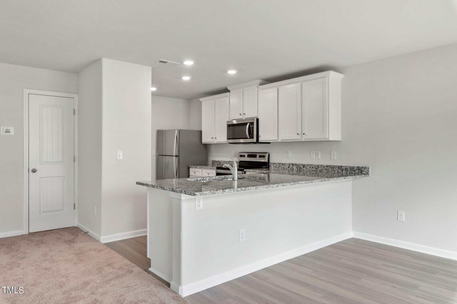 A modern kitchen with light-colored walls, a mix of carpet and wood flooring. It features stainless steel appliances, including a refrigerator and microwave, white cabinets, and granite countertops with recessed lighting.