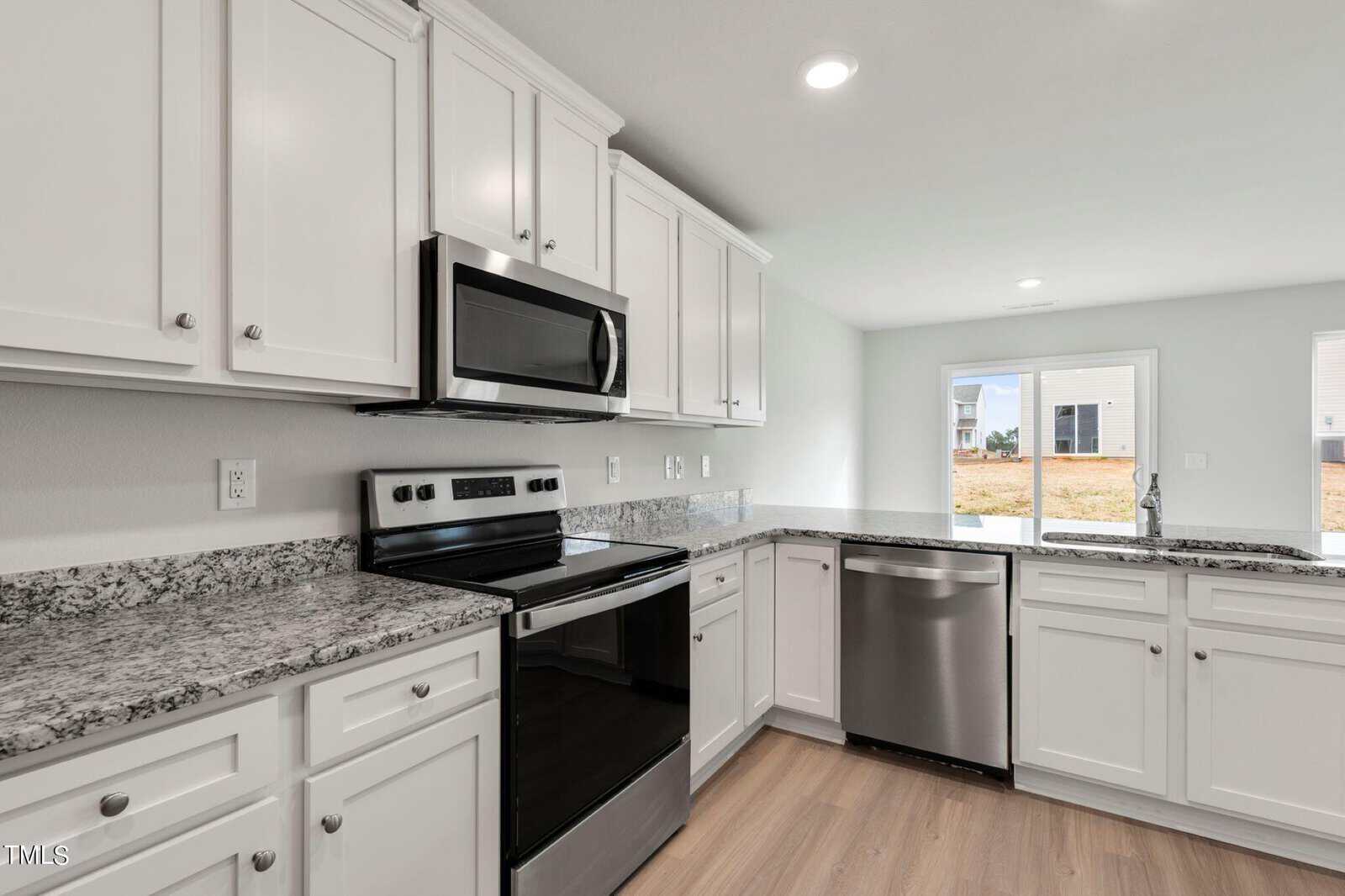 Modern kitchen with white cabinets, granite countertops, and stainless steel appliances, including an oven, microwave, and dishwasher. A large window provides natural light, and the floor is light wood.
