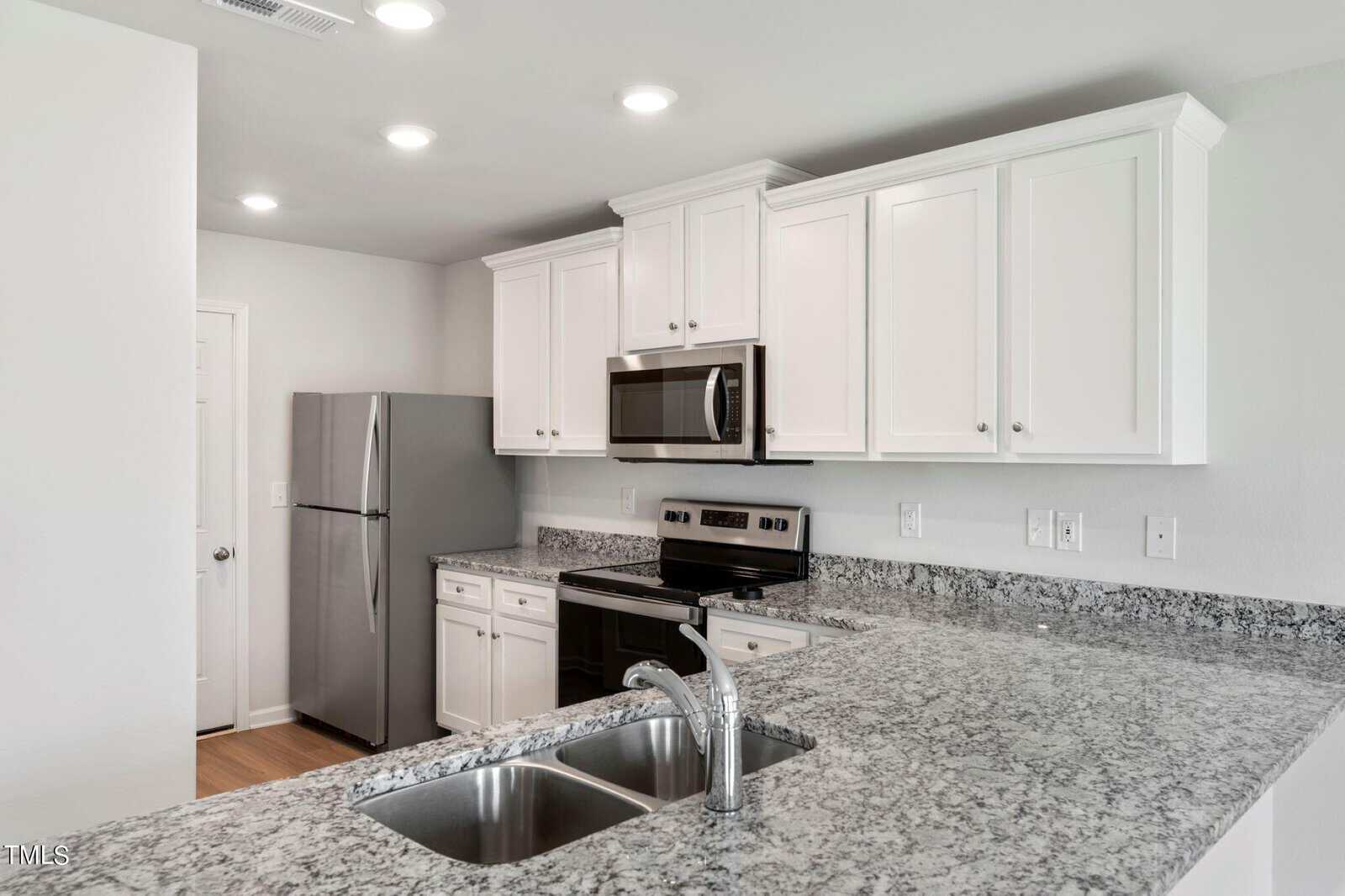 Modern kitchen with white cabinets, stainless steel appliances including a fridge and oven, and a granite countertop with a double sink. Recessed lighting brightens the space. The walls are painted white.