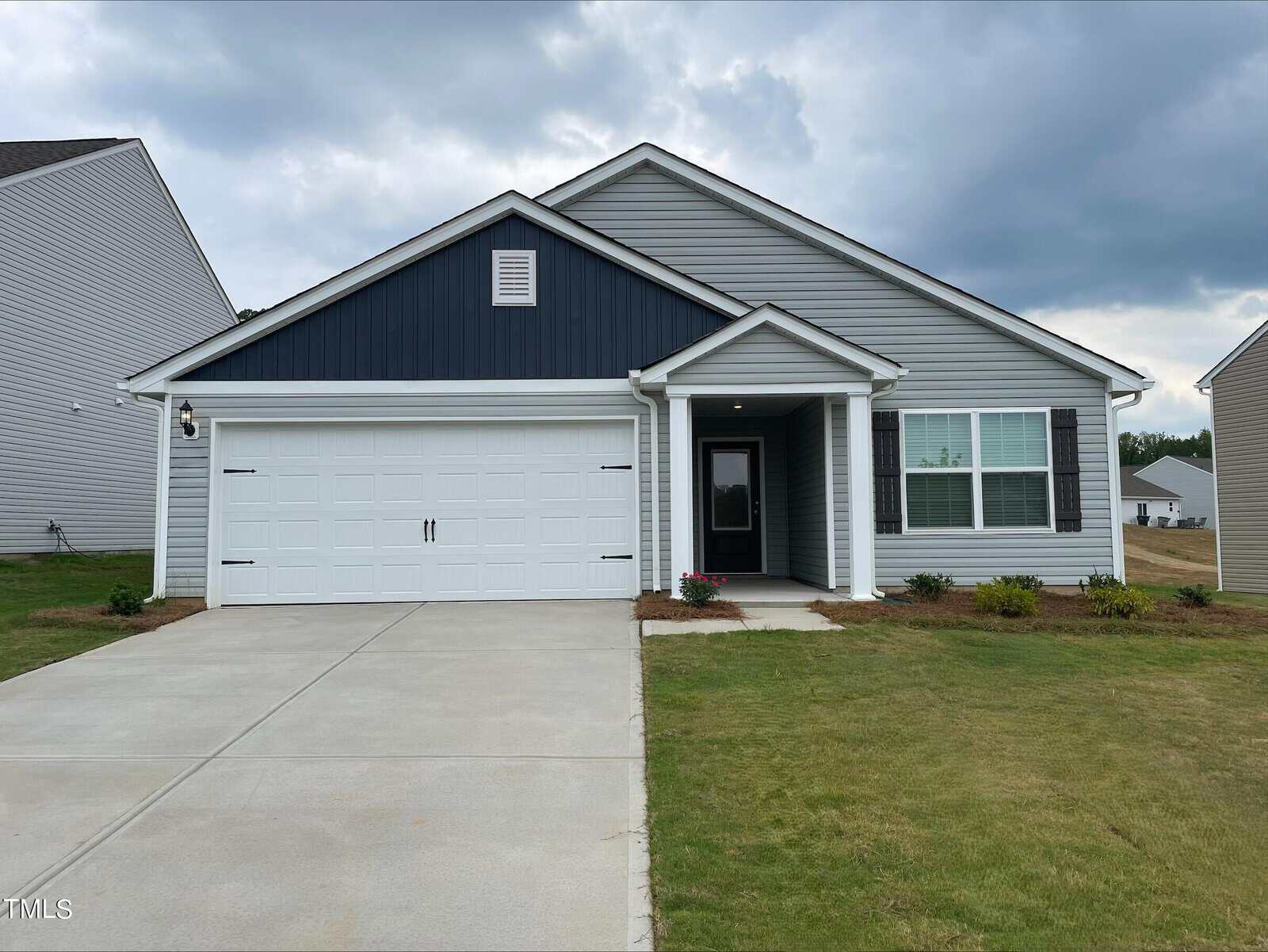 A single-story house with gray siding and dark blue gable, a white double garage door, covered front porch, and a concrete driveway. The yard has grass, small bushes, and flower beds. The sky is cloudy.