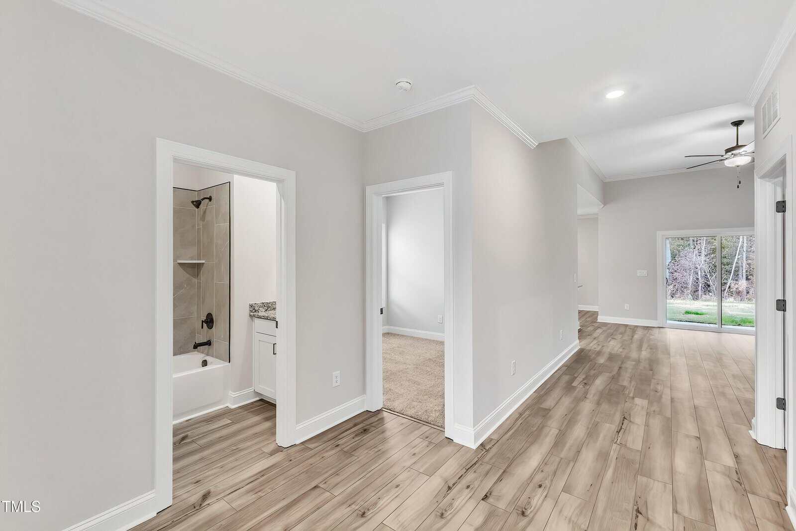 A hallway in a modern home features light-colored walls, wood-look flooring, an open living area with a ceiling fan, a bedroom with carpet, and a bathroom with a bathtub and granite countertop. Sliding doors lead outside.