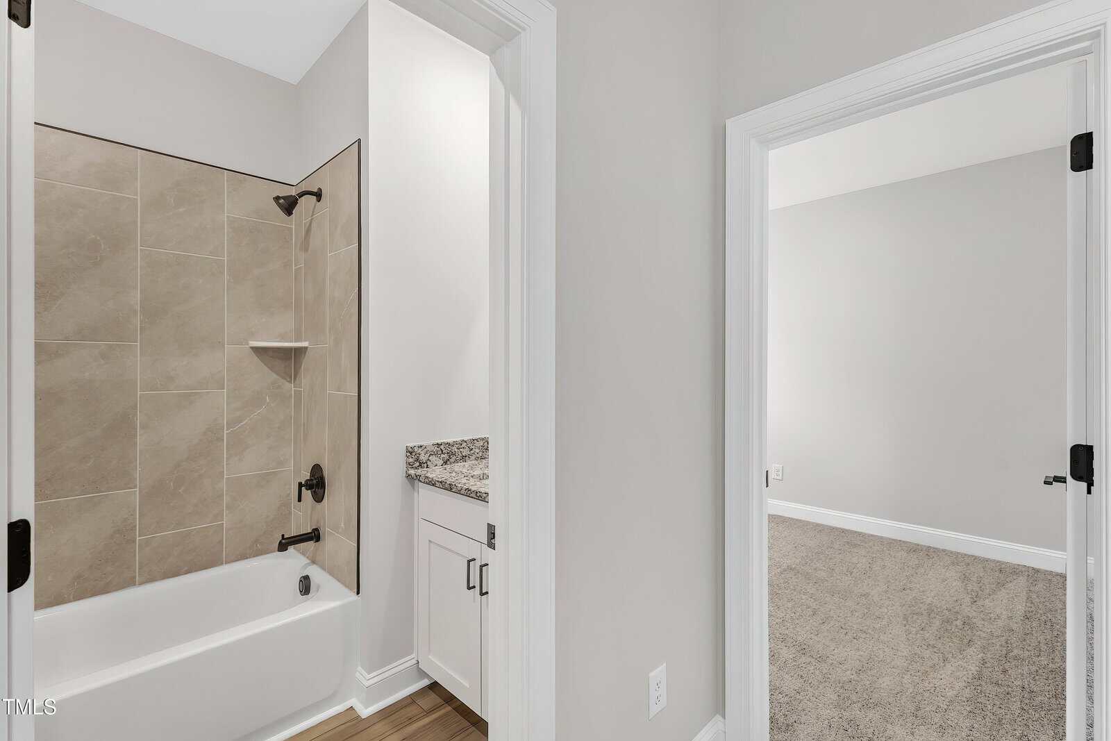 A bathroom with a bathtub and shower combination featuring beige tiles, a granite countertop with a cabinet, and an adjacent doorway leading to a carpeted room with light-colored walls.
