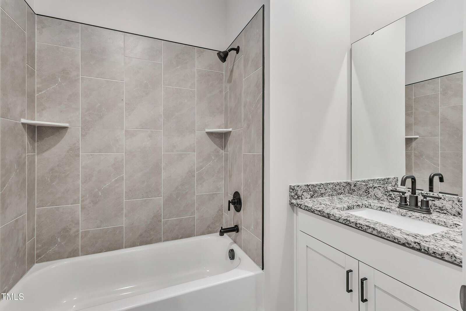 A bathroom with a bathtub and shower combination, beige wall tiles, two corner shelves, black shower fixtures, a white vanity with a granite countertop, and a large mirror above the sink.