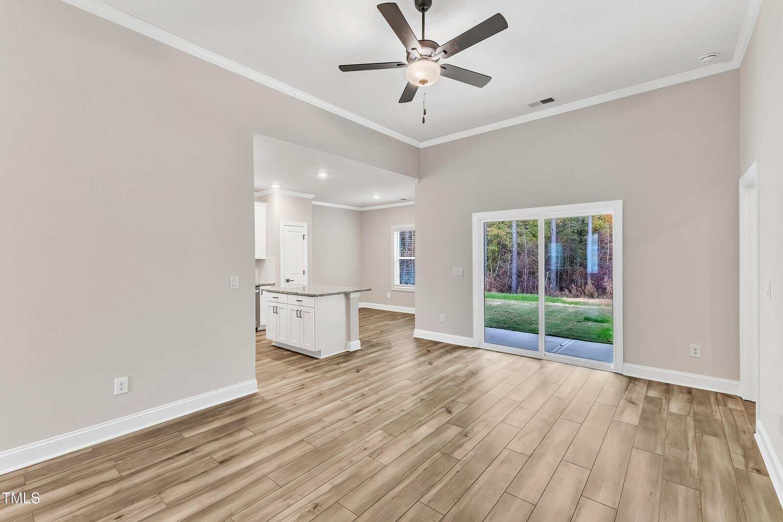 A spacious, empty living area with light wood flooring, beige walls, a ceiling fan, and sliding glass doors leading to a patio. The room connects to a white kitchen with an island and natural light from windows.