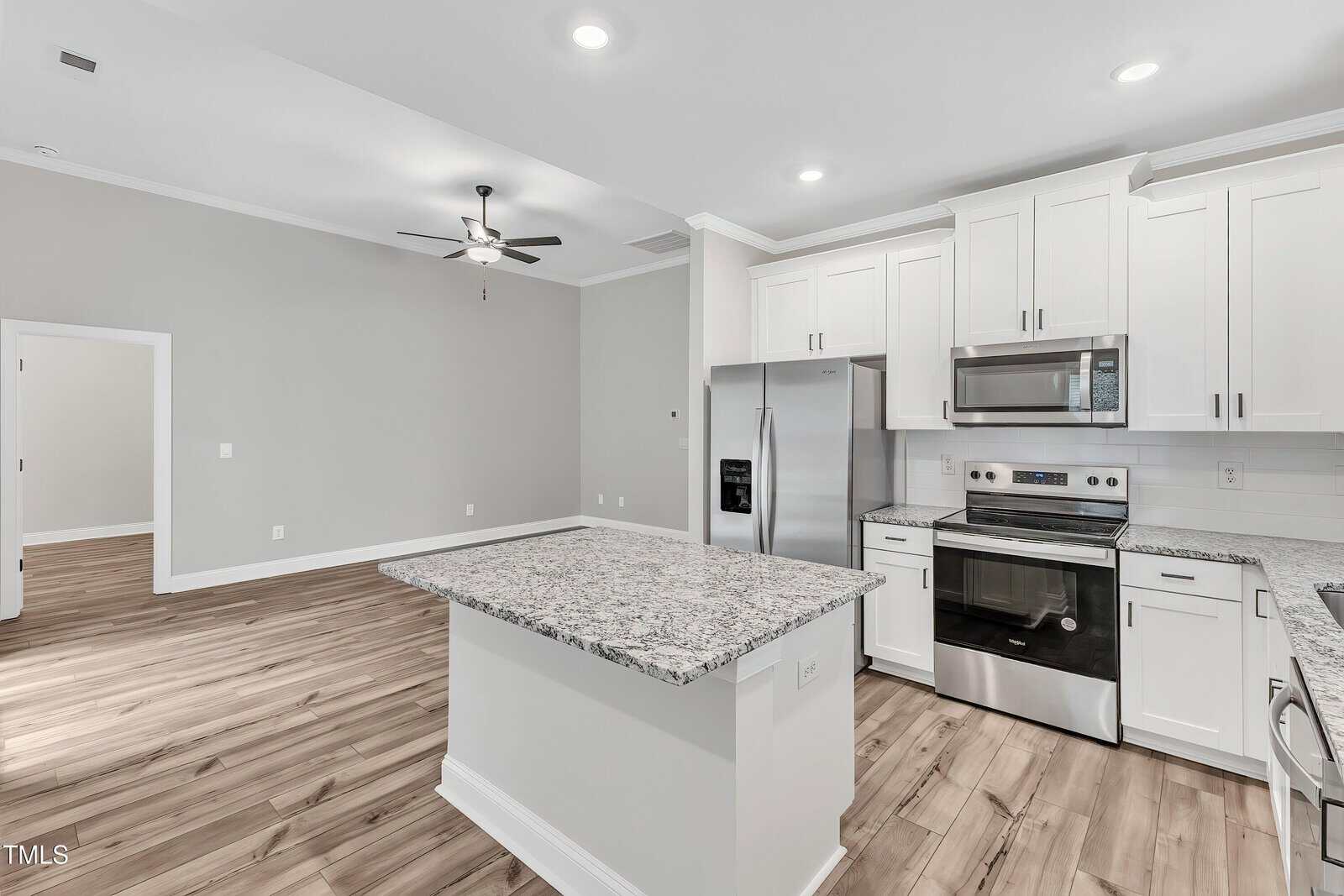 Modern kitchen with white cabinets, stainless steel appliances, granite countertops, and an island. The space has light wood flooring and opens to a living area with a ceiling fan and neutral-colored walls.