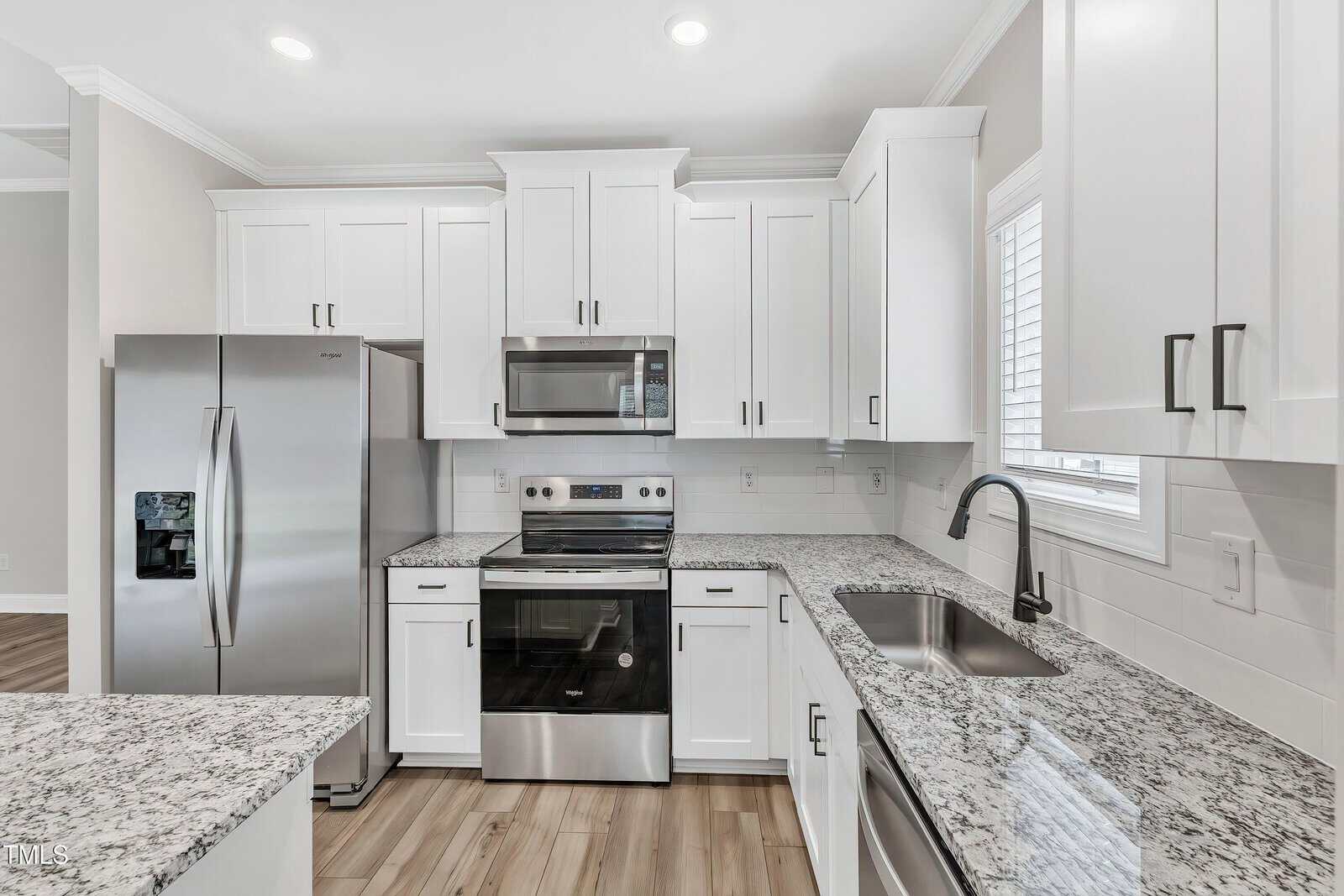 Modern kitchen with white cabinets, stainless steel appliances including a double-door refrigerator, microwave, and oven, granite countertops, an under-mount sink, and a window above the sink. Light wood flooring throughout.