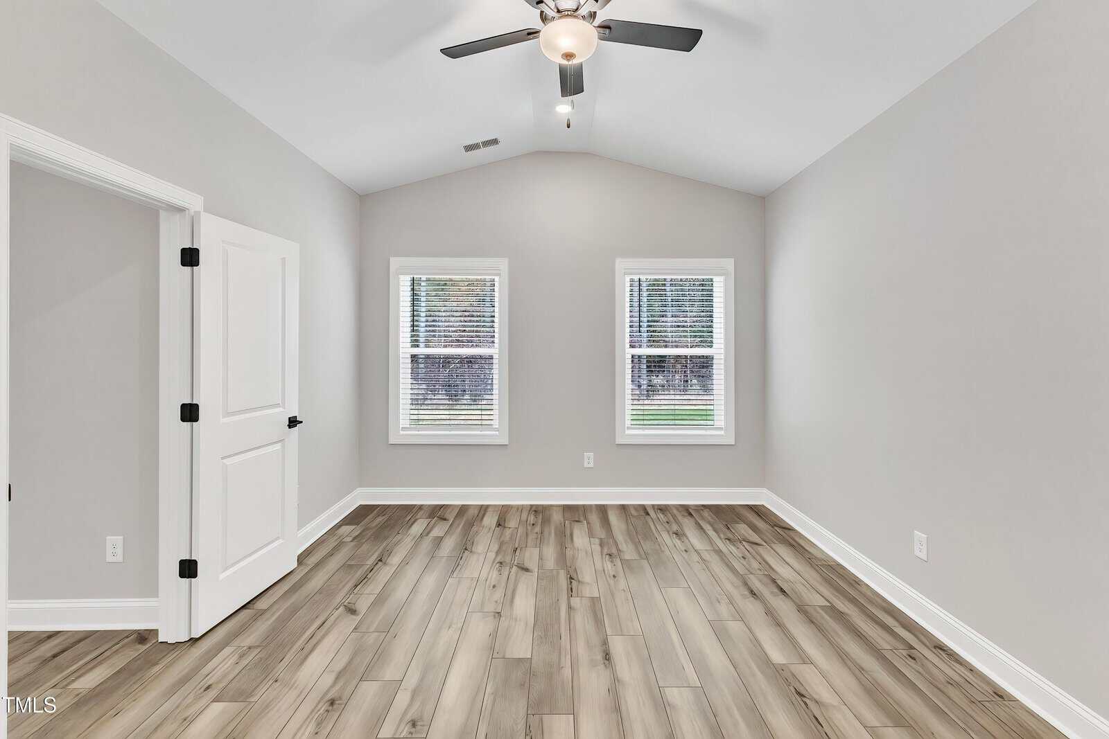 Empty room with light wood flooring, light gray walls, two windows with blinds, white trim, a ceiling fan with lights, and an open white double door on the left.