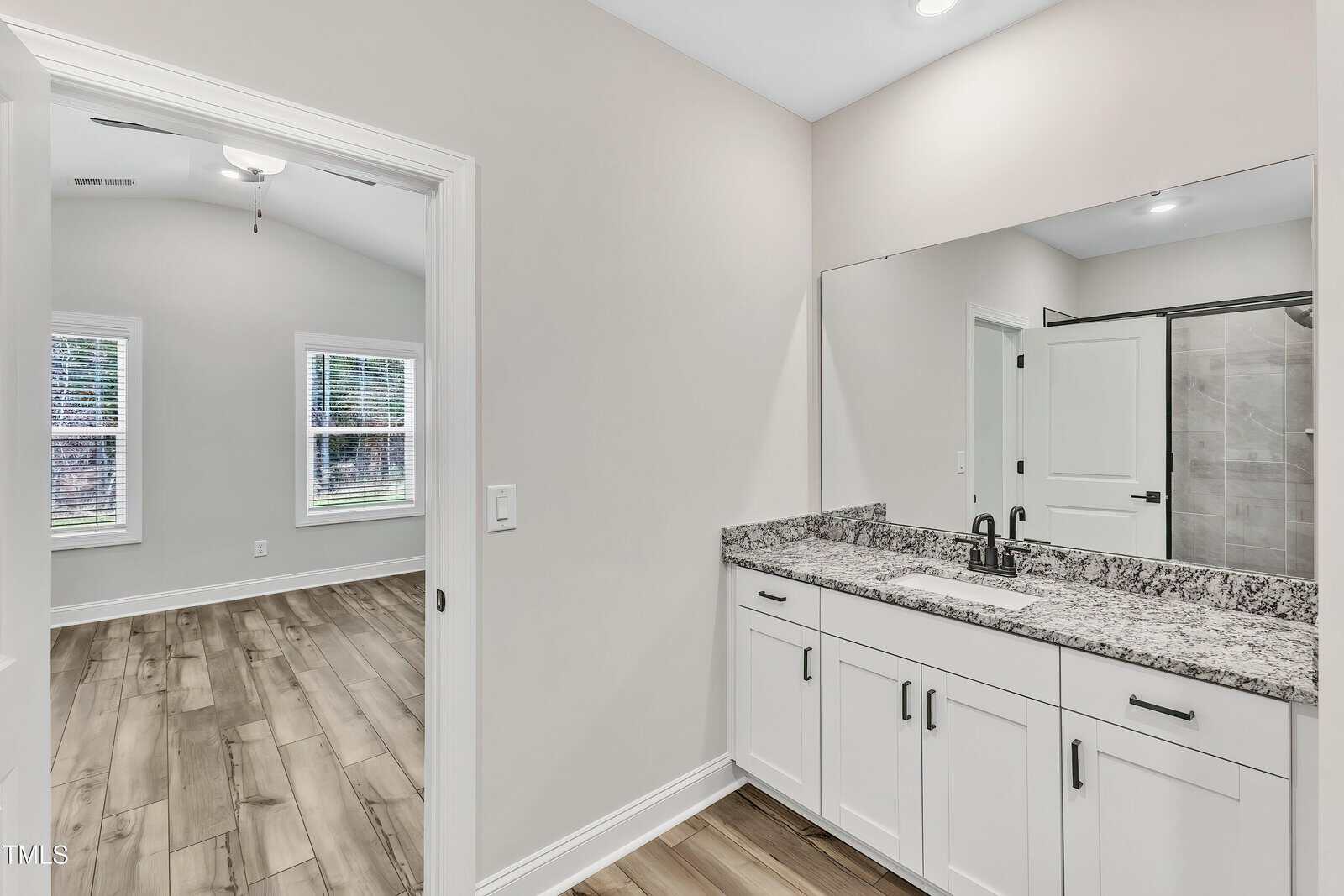 A modern bathroom with a granite countertop, white cabinets, and a large mirror, opening into a bedroom with wood-style flooring and two windows.