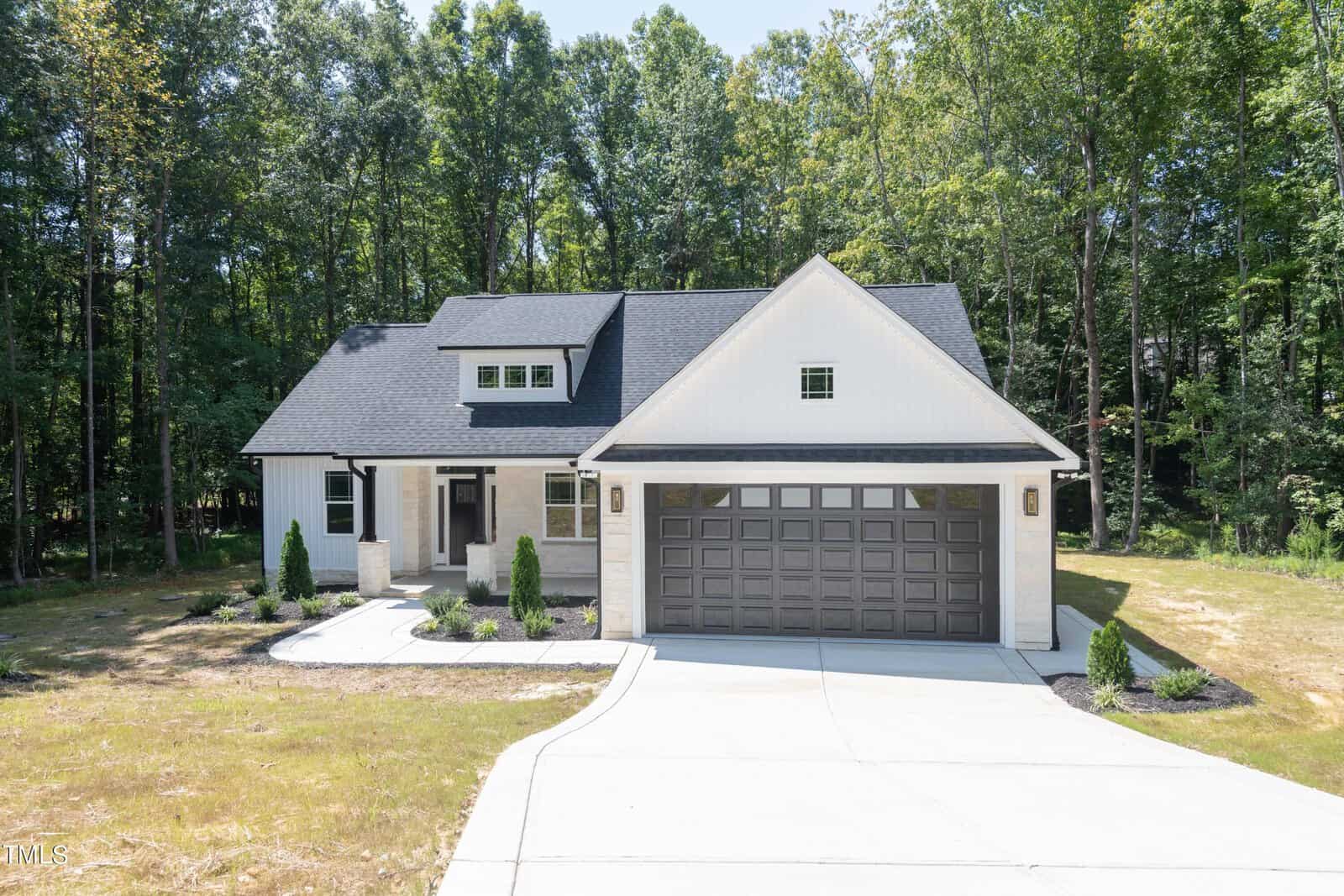 A modern, single-story house with white siding, a dark gray roof, and a double garage. The home is surrounded by a lawn, minimal landscaping, and tall trees in the background.
