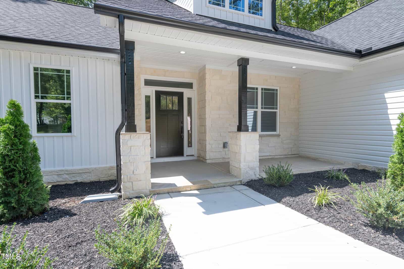 Front view of a modern house entryway with light stone accents, black columns, a glass-paneled front door, white siding, a covered porch, and neatly landscaped shrubs and plants.