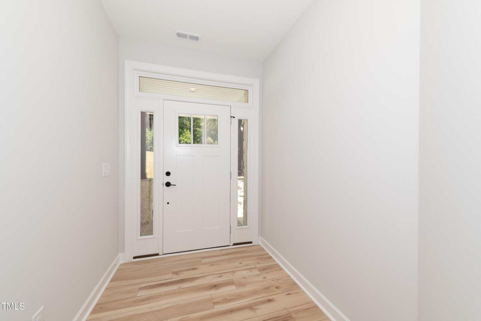 A bright, narrow entryway with light wood flooring and white walls, featuring a white front door with glass panels on either side and a transom window above, letting in natural light.