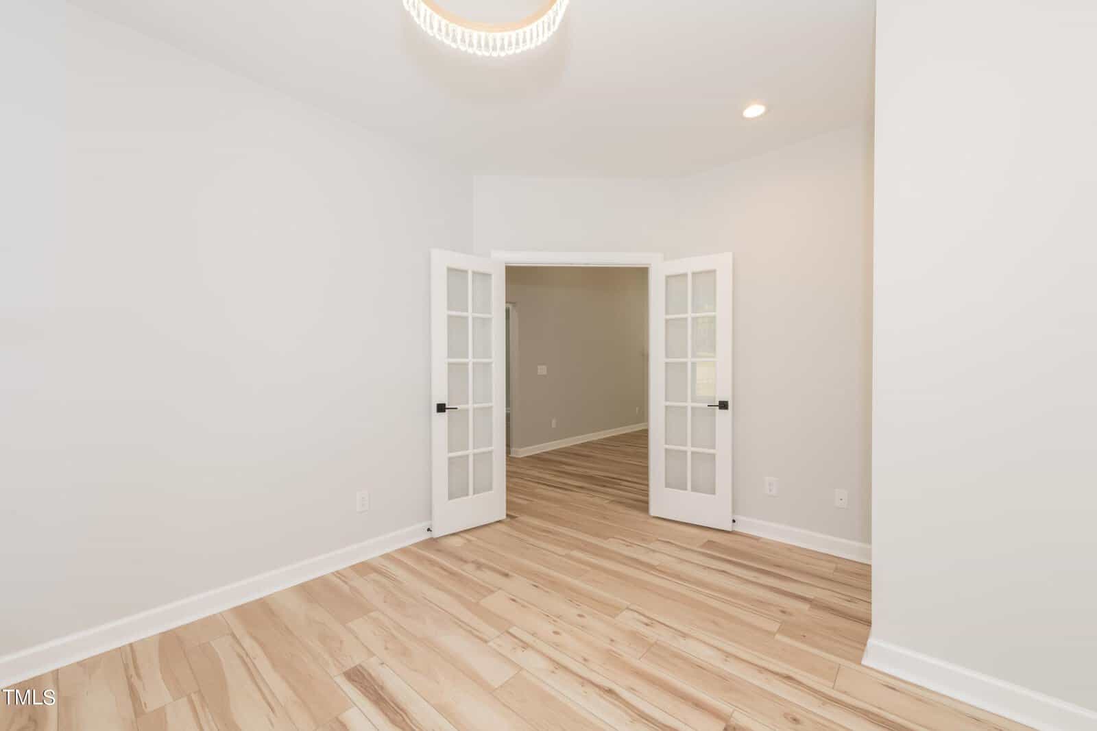 Bright, empty room with light wood floors, white walls, and glass French doors opening to another room with similar flooring. Ceiling light and recessed lighting are visible.